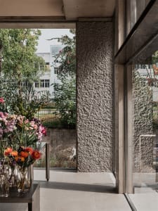 Modern lobby with concrete walls and large windows, featuring a vase of flowers and greenery.