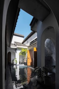 A minimalist residential hallway with concrete walls and floor, featuring an arched doorway leading to a courtyard with a pon