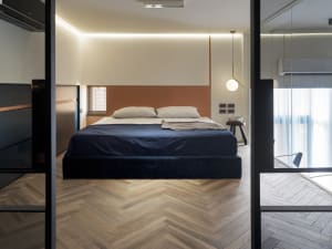 A modern minimalist bedroom with a wooden floor, fabric bedding, and a metal pendant light.