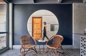 A circular window in a blue wall with a wooden door and woven chairs.