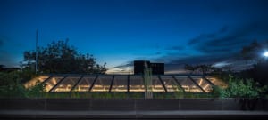 A modern greenhouse with a glass extension at dusk, featuring illuminated interior and dark blue sky with scattered clouds.