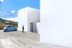 A modern white cube building made of concrete with a person walking by and a blue car parked nearby.
