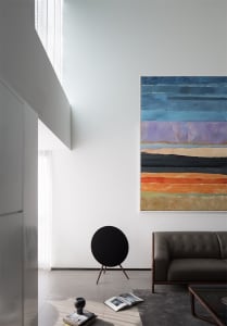 A modern living room featuring a large abstract painting on a wall, a round black speaker, a gray leather sofa, and a glass c