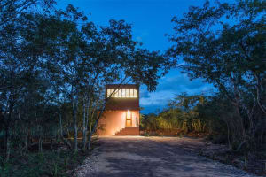 A small, clay-colored residential house with a lit entrance door and windows at dusk, surrounded by lush green vegetation.