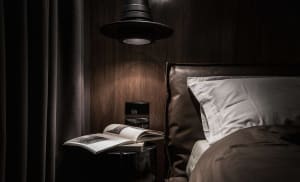 Dark, minimalist bedroom featuring a bed with a textile-leather headboard and a book on a side table with a pendant light abo