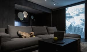 A cozy modern industrial living room with dark walls, a large dark sectional sofa, a coffee table with an apple, and a projec