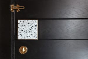 An elegant, black-colored wooden door with a geometric, white speckled terrazzo tile and a gold latch.