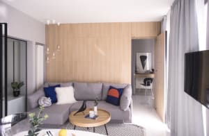 A modern minimalist living room featuring grey sofa with blue, orange, and white cushions, a wooden round coffee table, a met