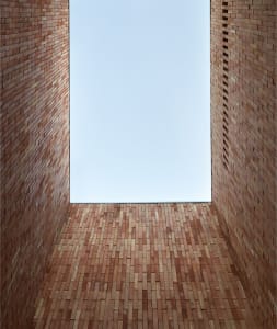 A rectangular room with brick walls and floor, looking up towards a blue sky through a window.