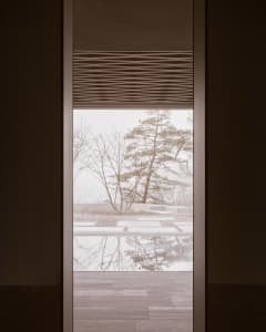 Metal blinds and a finished wooden floor frame a view of nature outside the window.