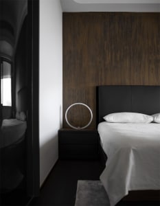 Modern minimalist bedroom featuring black leather headboard, wooden accent wall, and glass lamp on black nightstand.