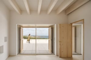 Minimalist room with open wooden doors leading to a patio, featuring a woman standing on the terrace overlooking the sea.