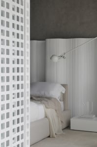 A minimalist bedroom featuring a white bed with a beige throw, a white lamp mounted on the wall, and a white perforated scree