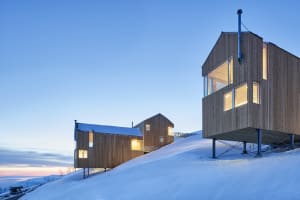 Modern wooden cabins with illuminated windows on a snowy slope at dusk or dawn.