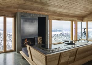 A modern living room with a wooden sectional sofa, glass tables, and a fireplace with a view of snowy mountains.