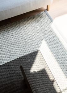 A close-up view of a modern grey knit rug with a sunlit pattern, placed under a grey cushioned sofa on a light wooden floor.