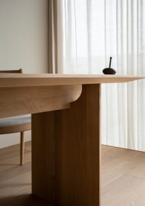 A minimalist dining room featuring a modern oak wood table with a unique curved design, adjacent to a window with sheer curta