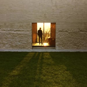 A man standing in a modern house with glass walls at sunset.
