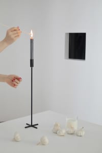 A white countertop with a black candle holder, a lit black candle, a glass of milk, and several garlic bulbs. A hand with red