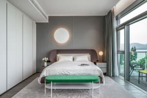 A modern bedroom with white furniture, a green ottoman, and a view of mountains outside.