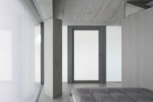 A minimalist bedroom with concrete walls, floor, and ceiling, featuring a bed with a gray cover.