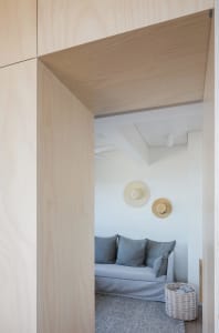 A minimalist living room with a white sofa, grey cushions, and a light wooden wall and floor.