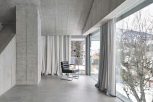 A minimalist living room with concrete walls, floor-to-ceiling windows, and a black armchair.