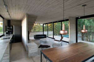 Modern kitchen with concrete walls, dining table, and living room with large windows and outdoor patio.