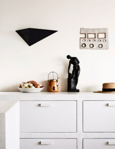 A minimalist kitchen with white cabinets, concrete countertops, a bowl of colorful vegetables, and a black sculpture on the c
