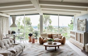 A modern living room with a tan leather sofa, round coffee table, and large windows overlooking greenery.