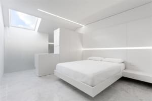 A modern white bedroom with a floating bed, white walls, and a white ceiling light.
