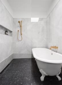 A minimalist bathroom with marble walls and porcelain tiles, featuring a clawfoot bathtub and a walk-in shower with gold fixt