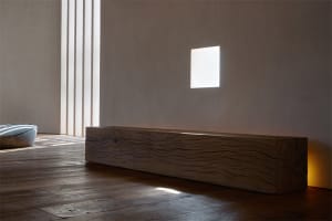 A minimalist interior space featuring a wooden log bench and geometric light patterns on a white wall.