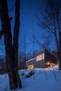 A modern cabin with wooden exterior and large windows against a snowy background at dusk.