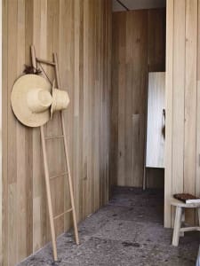 A minimalist interior with wooden walls, a stone floor, a rustic wooden ladder with straw hats, and a small wooden stool.
