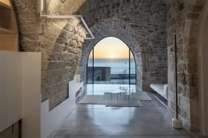 A minimalist living space featuring stone walls, concrete flooring, and glass doors leading to an ocean view.