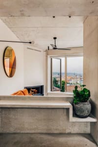 A modern living room with concrete walls, an orange couch, a fireplace, and a large window with a city view.