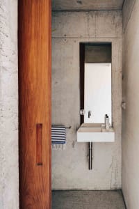 A minimalist bathroom with concrete walls, a wooden door, a white sink, and a striped hand towel hanging on a towel ring.