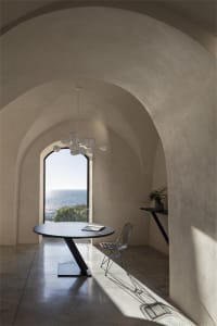 A minimalist interior design featuring concrete walls and floor, a circular table, and a large window overlooking the sea.