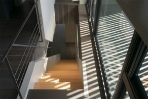A modern staircase with glass walls and a wooden floor, featuring a large window with vertical blinds.