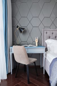 A minimalist bedroom featuring a workspace with a blue desk, beige chair, black table lamp, and a hexagonal patterned wall, a