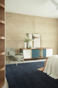 A minimalist bedroom featuring a wooden storage unit, a fabric armchair, and a navy blue rug.