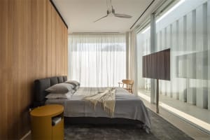 A modern minimalist bedroom featuring a wooden wall and floor, large glass windows, and a comfortable bed with grey bedding a