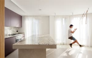 A modern kitchen features a large granite marble countertop and island with purple cabinets and white curtains in the backgro