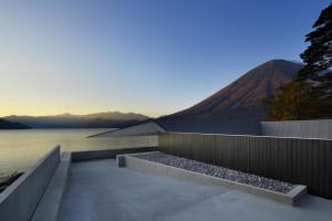 A modern concrete residence with a rooftop terrace overlooking a lake and mountains.