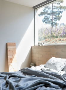 A modern bedroom with a concrete bed frame and large window overlooking a garden.