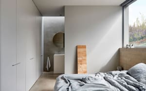 Minimalist bedroom with concrete walls and floor, featuring a bed and a small sink with a circular mirror.