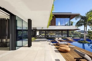 Modern residential exterior featuring wood, glass, and concrete materials, with a large pool and outdoor seating area.