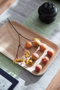 A wooden tray with small, round, yellow and pink fruits, a yellow flower stem, and a ceramic incense burner on a green and gr