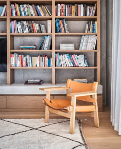 A modern library interior with a wooden framed bookshelf filled with books, and a comfortable leather armchair with wooden le